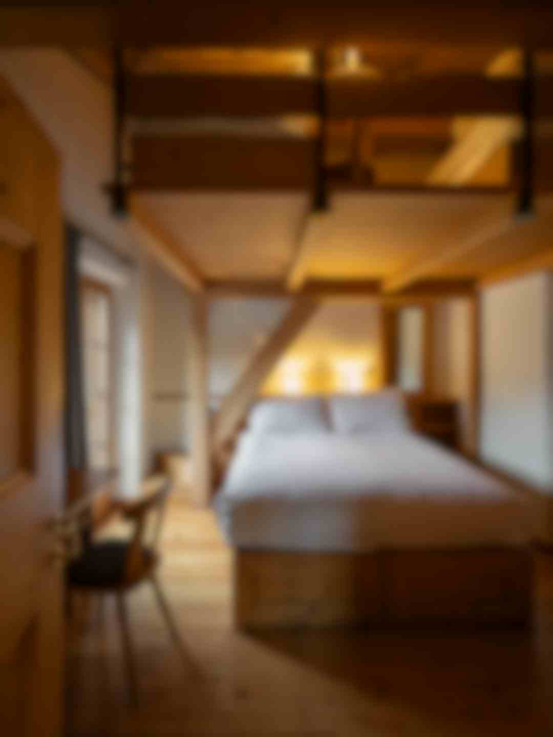 Camera in stile alpino con letto matrimoniale incorniciato da travi in legno a vista. Pavimento e arredi in legno chiaro, scrivania vicino alla finestra e illuminazione soffusa che rende l’ambiente accogliente
