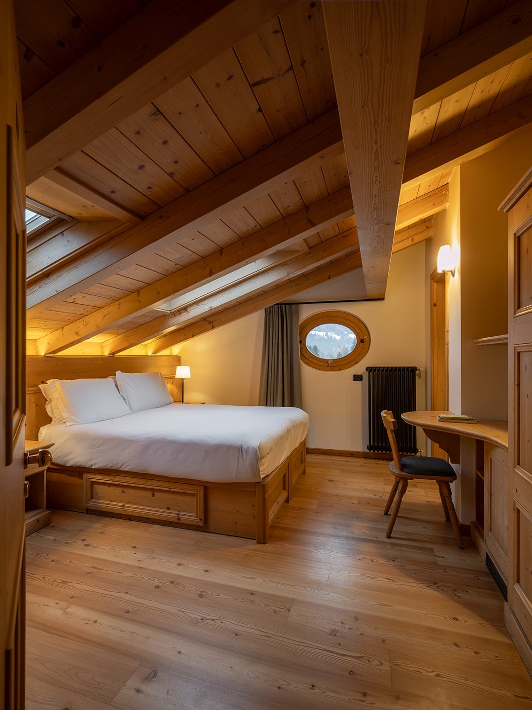 Camera mansardata in stile alpino a Bormio con letto matrimoniale, travi in legno a vista e finestra rotonda con vista sulle montagne. Scrivania in legno e luce calda rendono l’ambiente accogliente e raccolto.