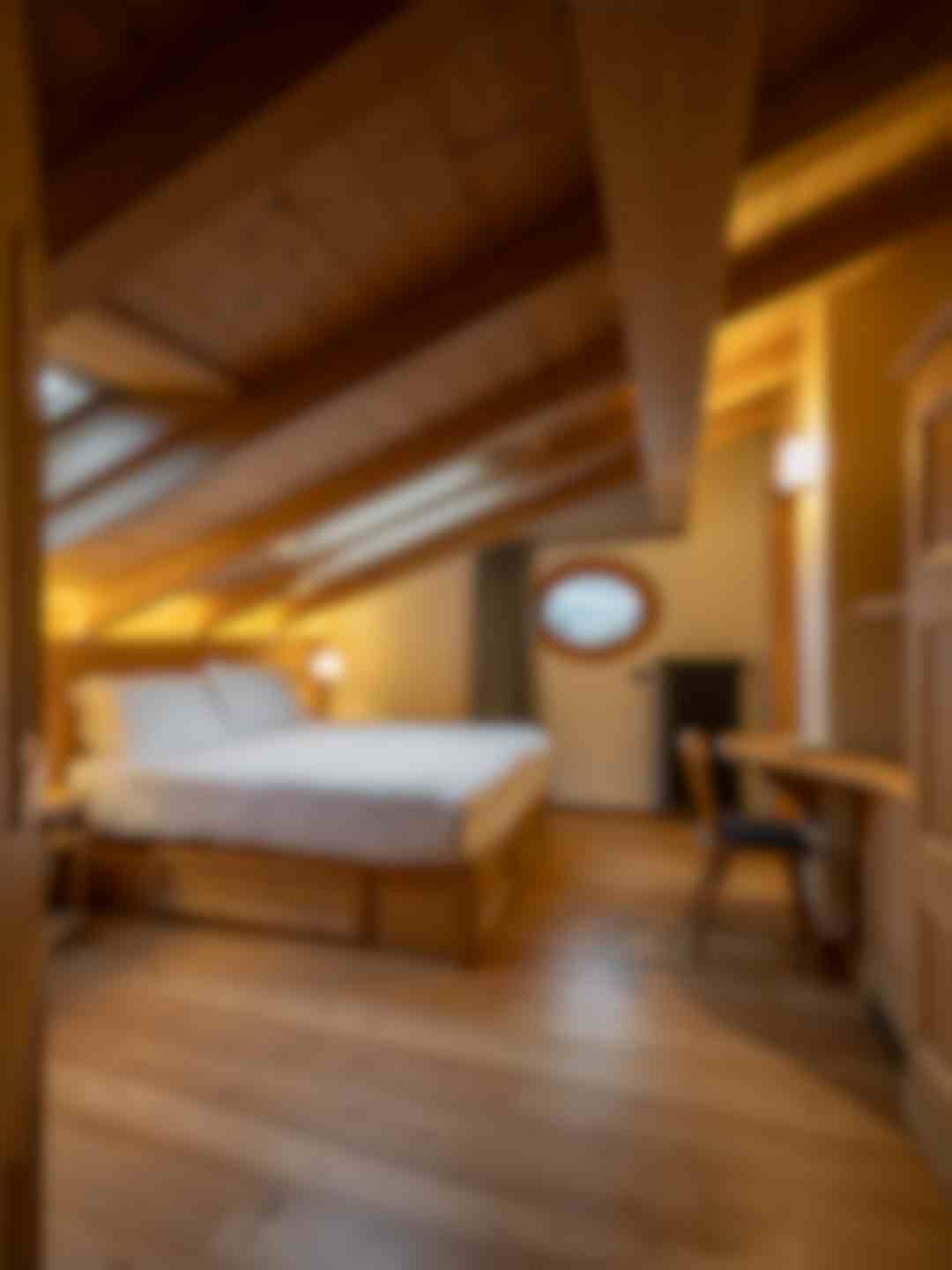 Camera mansardata in stile alpino a Bormio con letto matrimoniale, travi in legno a vista e finestra rotonda con vista sulle montagne. Scrivania in legno e luce calda rendono l’ambiente accogliente e raccolto.