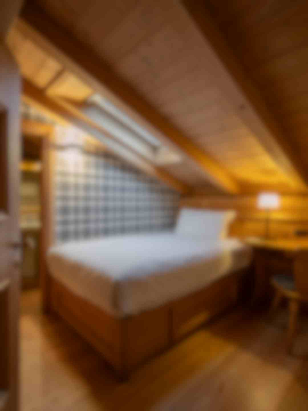 Camera mansardata in legno in stile Alpi&Golf Hotel con letto matrimoniale, parete rivestita in tessuto tartan e finestra sul tetto. Illuminazione soffusa e arredi in legno chiaro creano un’atmosfera calda e intima.
