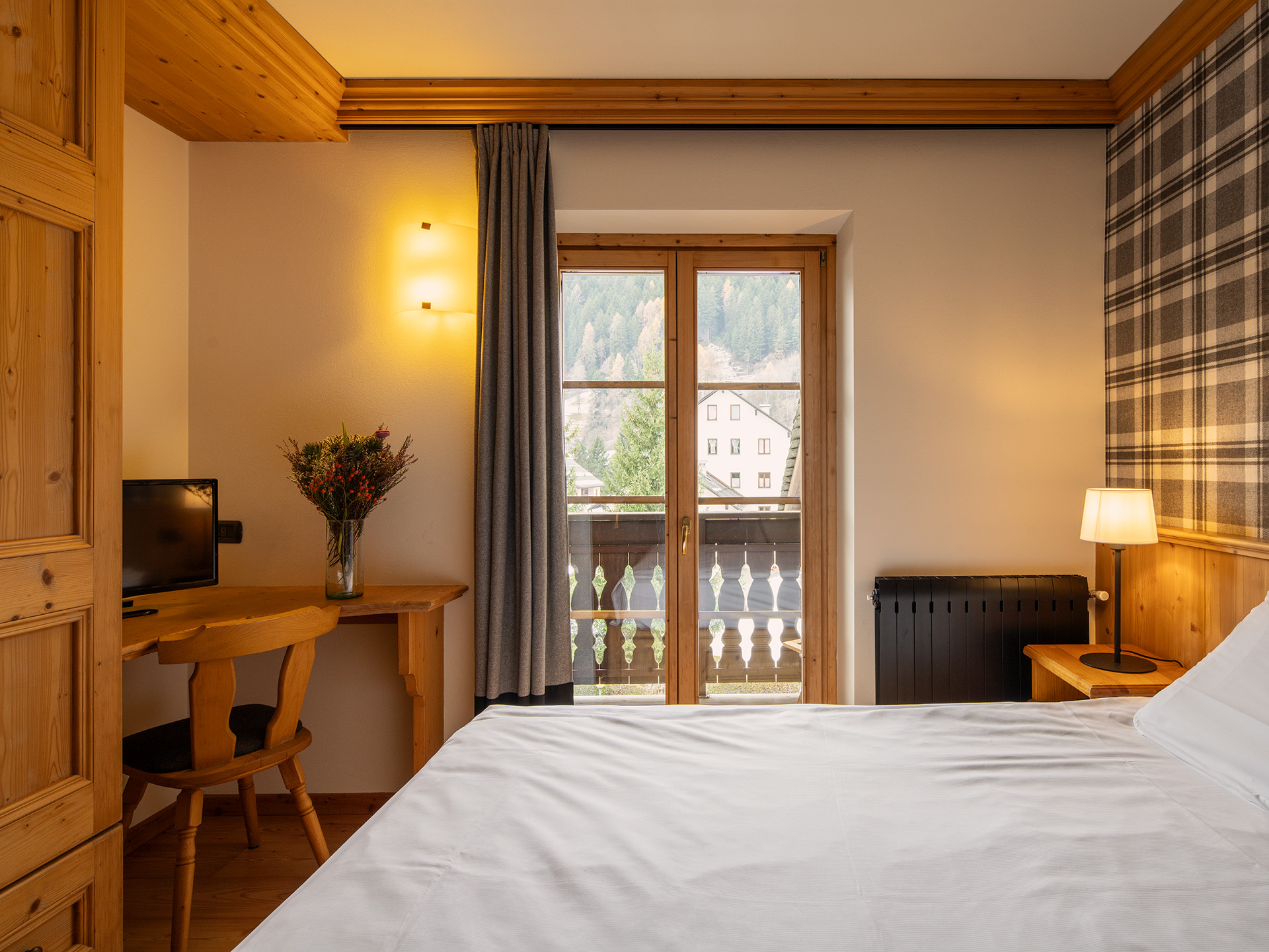 Stanza in legno e carta da parati con motivi tartan, letto matrimoniale con lenzuola bianche, scrivania con vaso di fiori e tv. Finestra vista montagne.