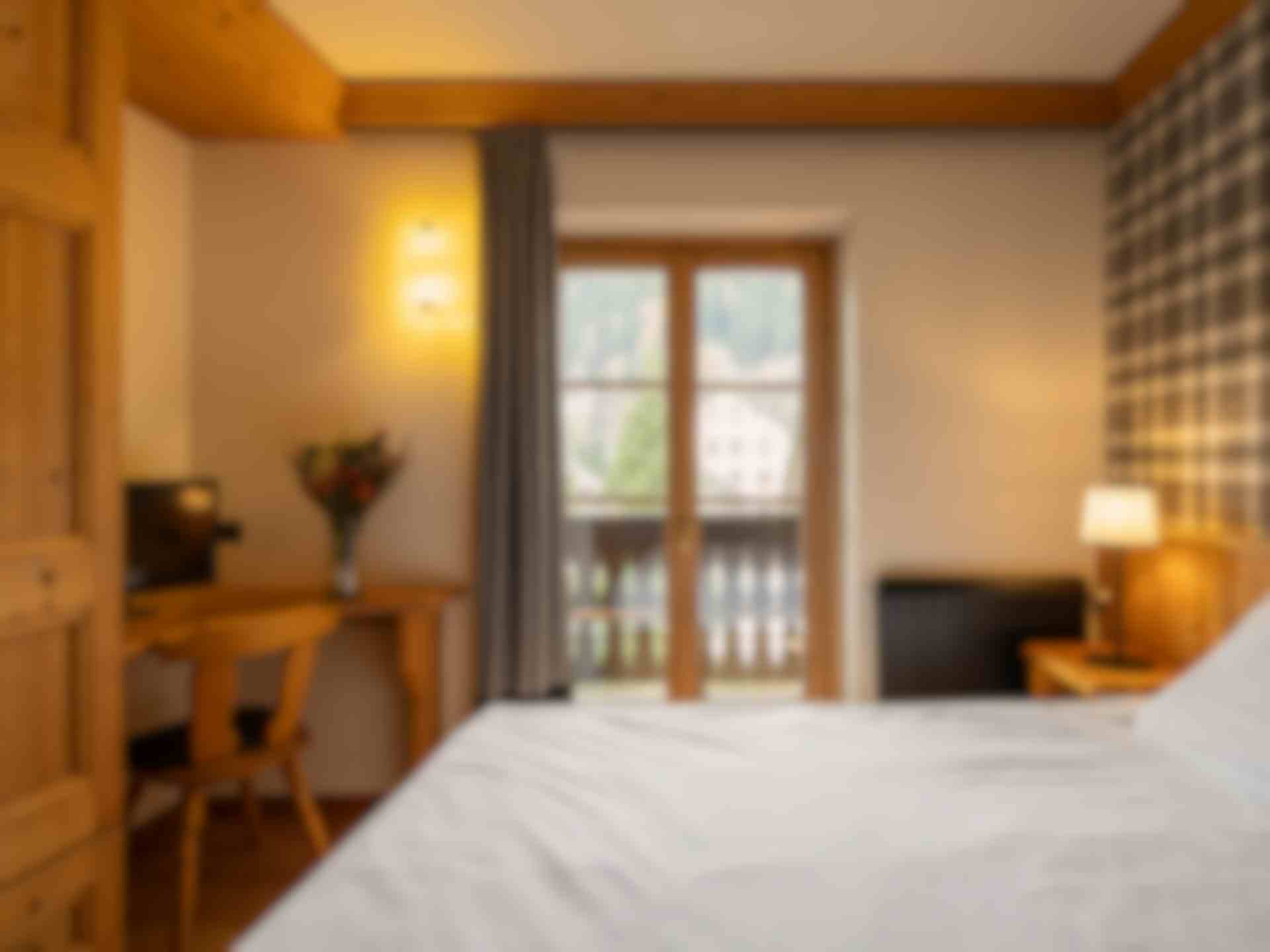 Stanza in legno e carta da parati con motivi tartan, letto matrimoniale con lenzuola bianche, scrivania con vaso di fiori e tv. Finestra vista montagne.