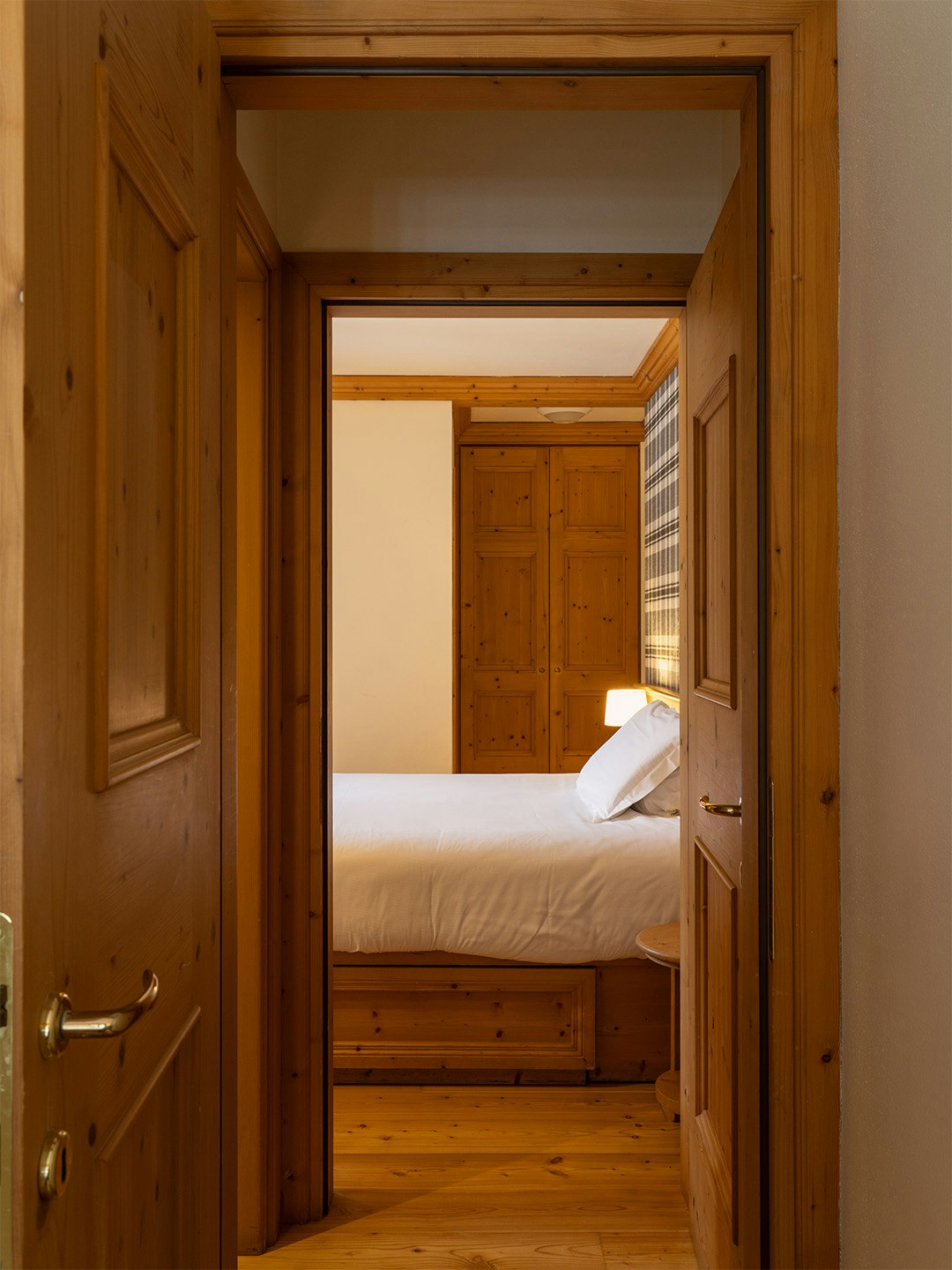 Porta aperta su camera da letto principale, letto matrimoniale con lenzuola bianche e doppi cuscini, porte e armadi in legno di abete.