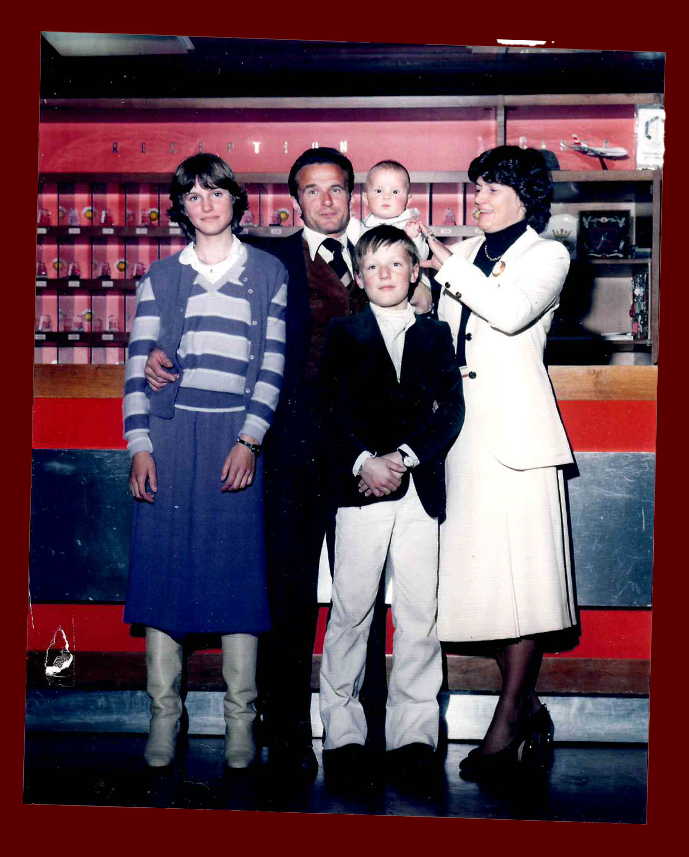 Famiglia con tre figli davanti alla reception dell'hotel, atmosfera anni '80 con arredamento rosso