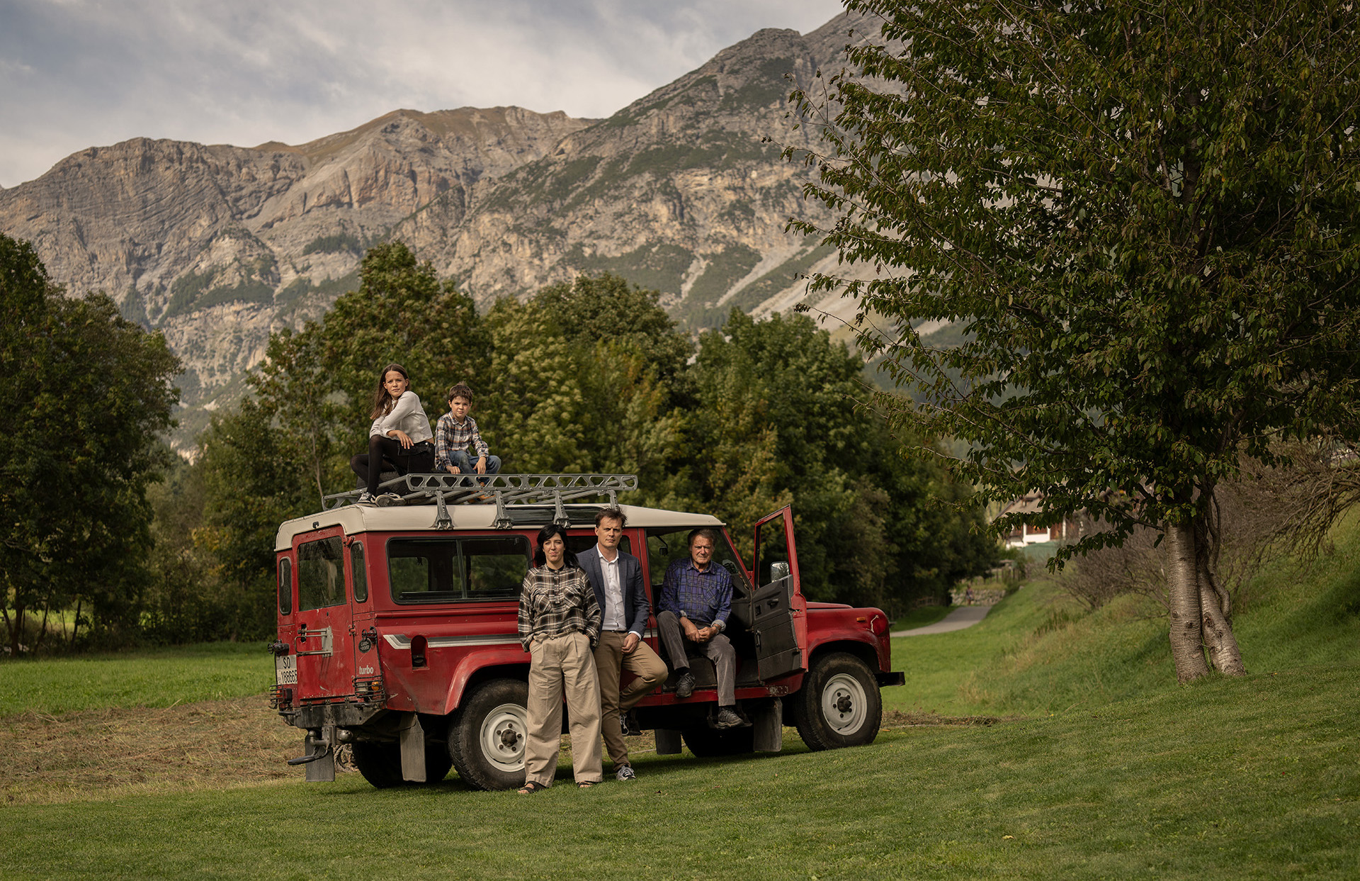 Famiglia dell’Hotel Alpi & Golf a Bormio riunita accanto a una Land Rover rossa d’epoca, con le montagne alpine sullo sfondo e prati verdi intorno.