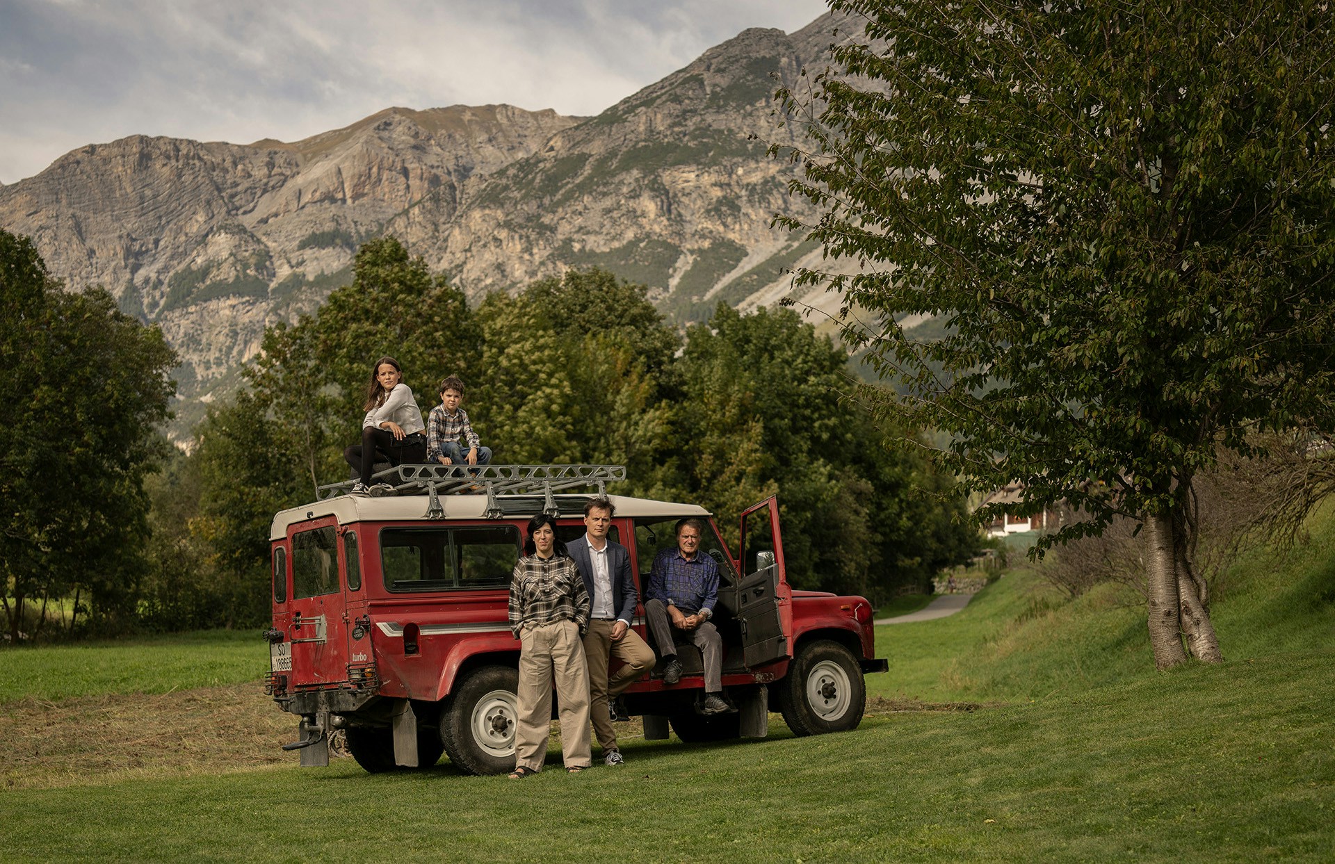 Famiglia dell’Hotel Alpi & Golf a Bormio riunita accanto a una Land Rover rossa d’epoca, con le montagne alpine sullo sfondo e prati verdi intorno.