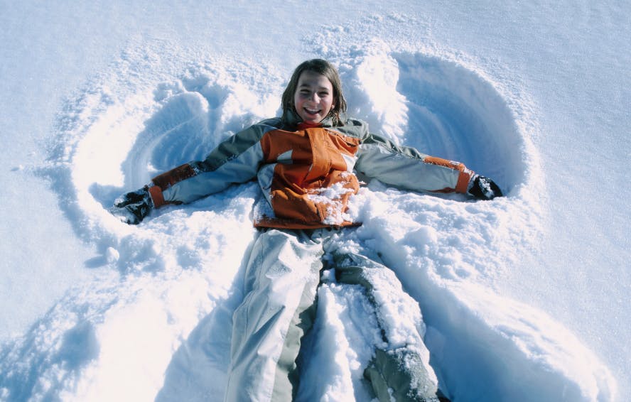 alt=young girl smiling in orange jacket doing a snow angel and having fun