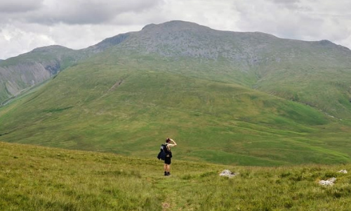A wide shot of a man walking though a green landscape with rocky hills