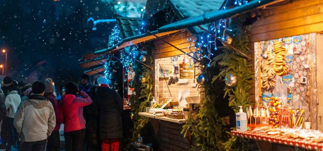 A shot of Christmas market stalls, and warmly-dressed customers mingling around the stalls. Featuring pine decorations and sparking lights. 