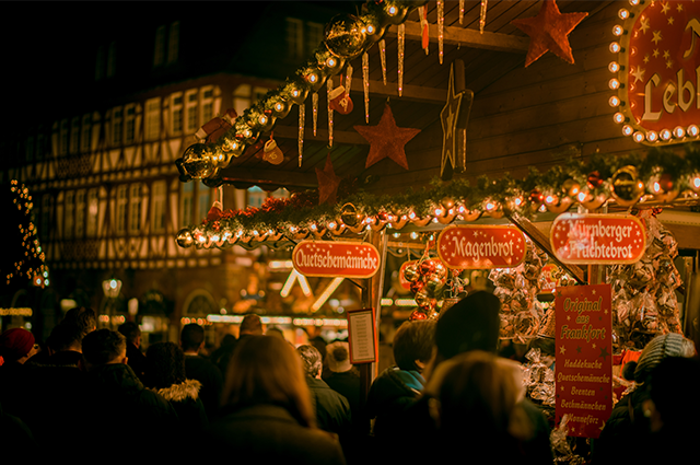 Visitors to a traditional German Christmas market with warm fairy lights lighting the area and Christmas decorations hanging from the chalet-style building 