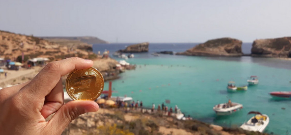 A photo of the coastline somewhere in Greece with turquoise seas. In the forefront, someone is holding a Cryptocurrency coin in front of the camera