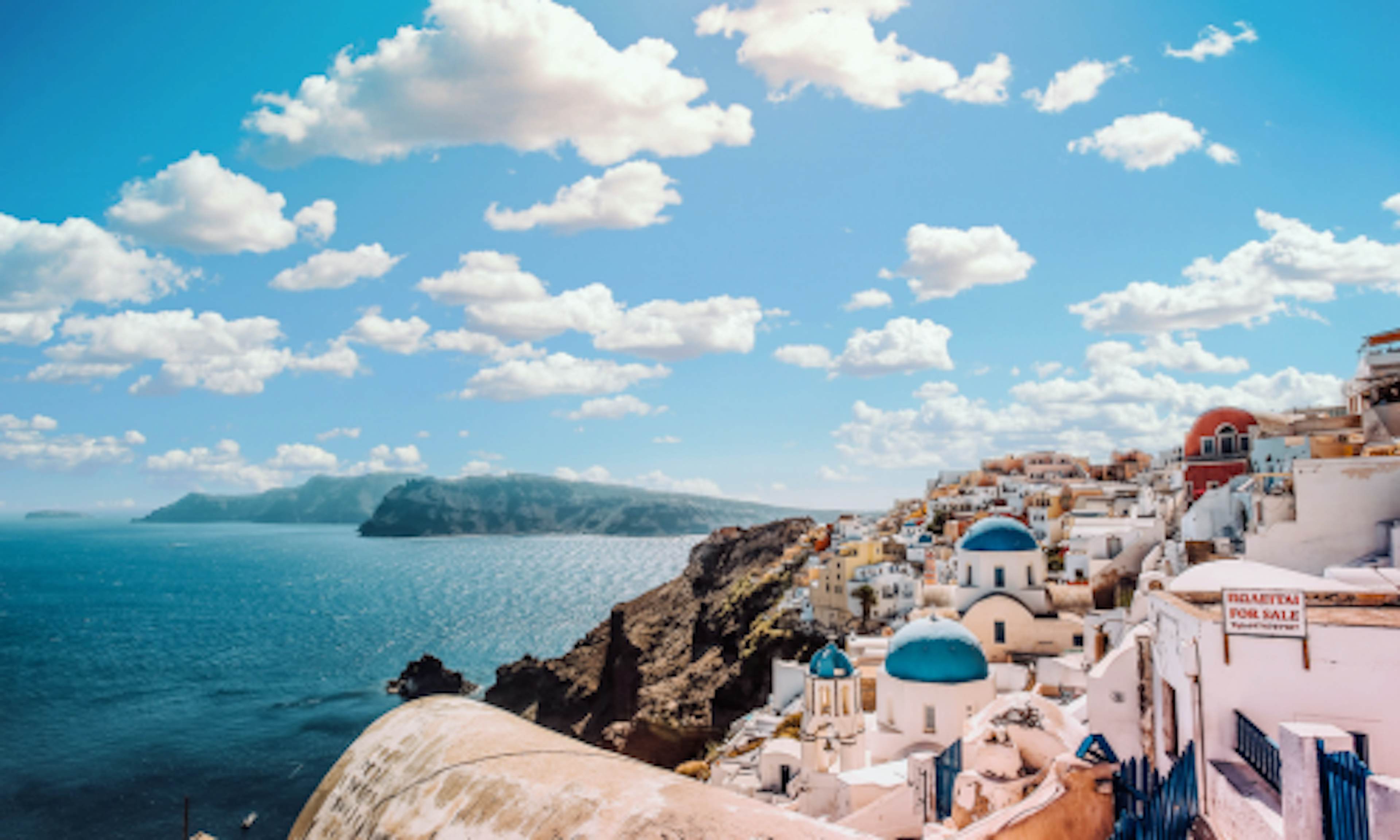 Santorini, Greece. White washed houses on side of hill, blue roofs. Sunny skies with white clouds.