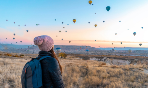 A woman staring up at hot air balloons at sunset over a desert landscape 