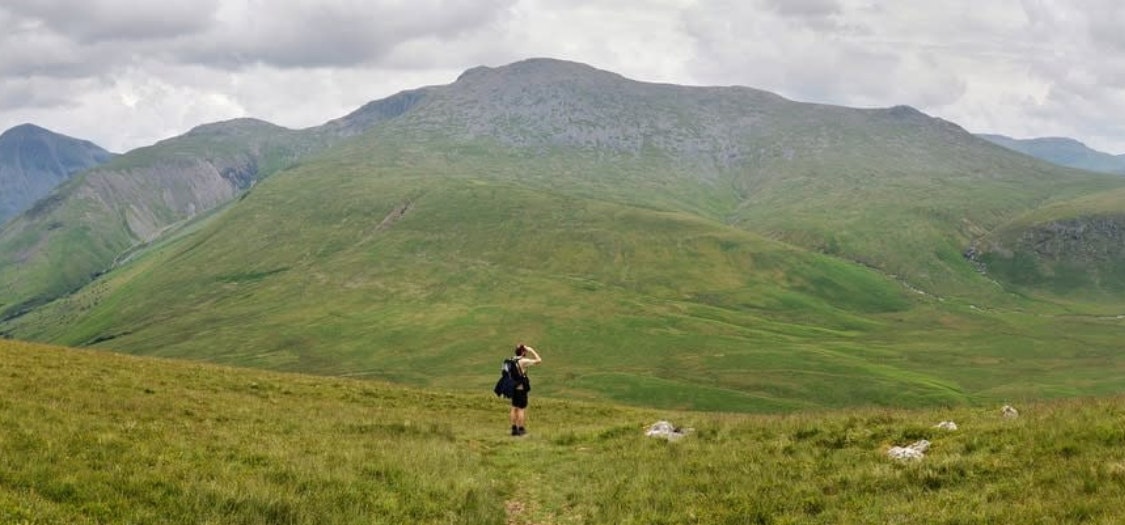 A wide shot of a man walking though a green landscape with rocky hills