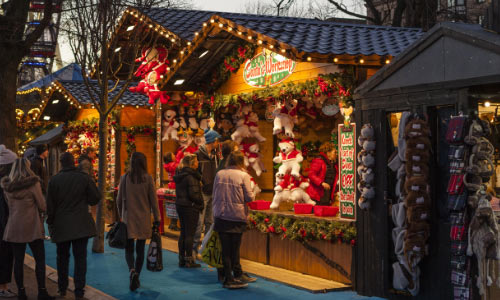 A shot of Christmas market stalls, and warmly-dressed customers mingling around the stalls. Featuring pine decorations and sparking lights. 