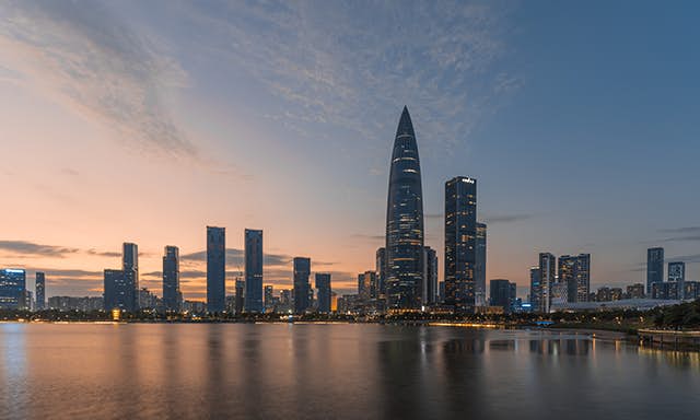 The futuristic skyline of Shenzhen bathed in dim glow from the setting sun casting reflections over the water below