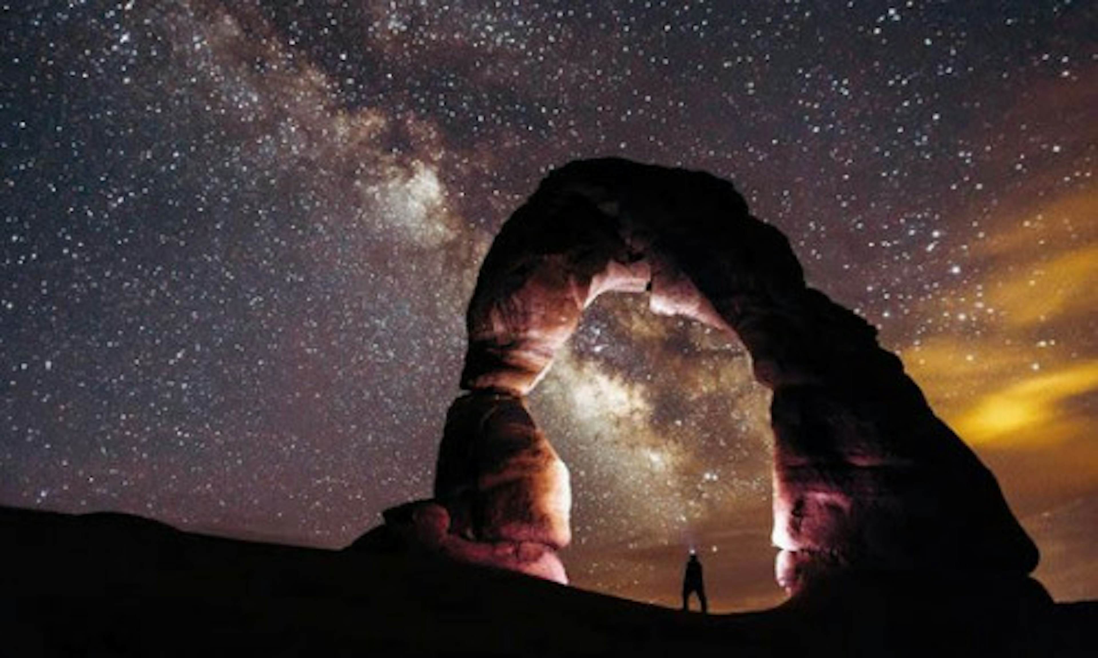 A small silhouette of a stargazer looking up through a stone arch in the Arches National Park, with a brilliant view of the stars above