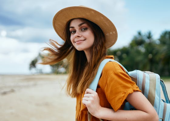 A happy woman with a backpack and sunhat walking along a beach, looking back at the camera and smiling 
