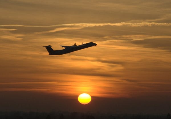 A twin-propeller aircraft flying through the sky lit up by the orange setting sun