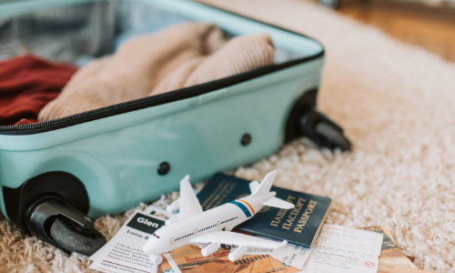 A low shot of an open teal suitcase on a shaggy cream rug. There's items starting to be packed, including a red jumper. Travel documents including a passport and small model of a plane are in front 