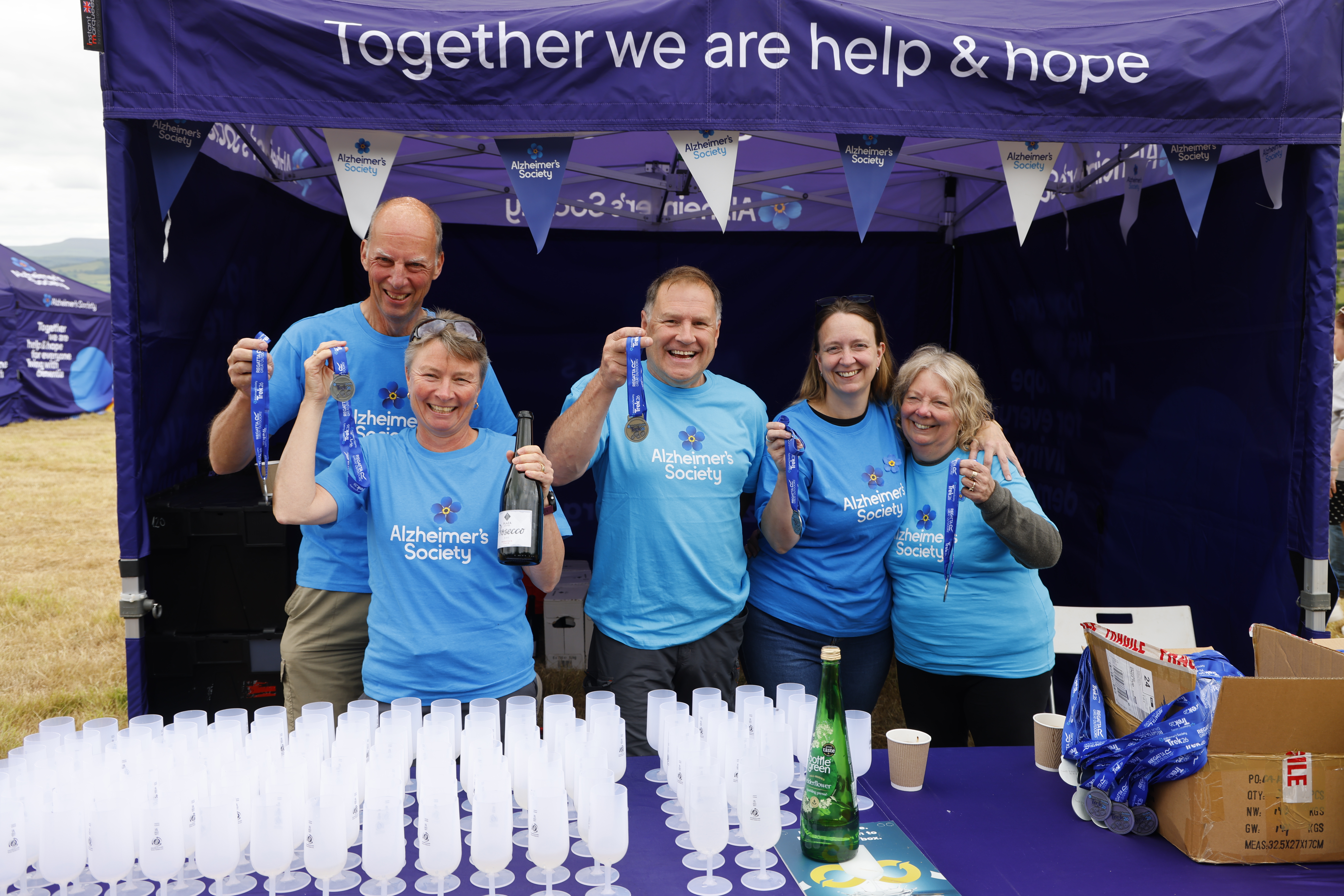 Five Trek26 volunteers standing behind a table filled with prosecco flutes and holding up medals.