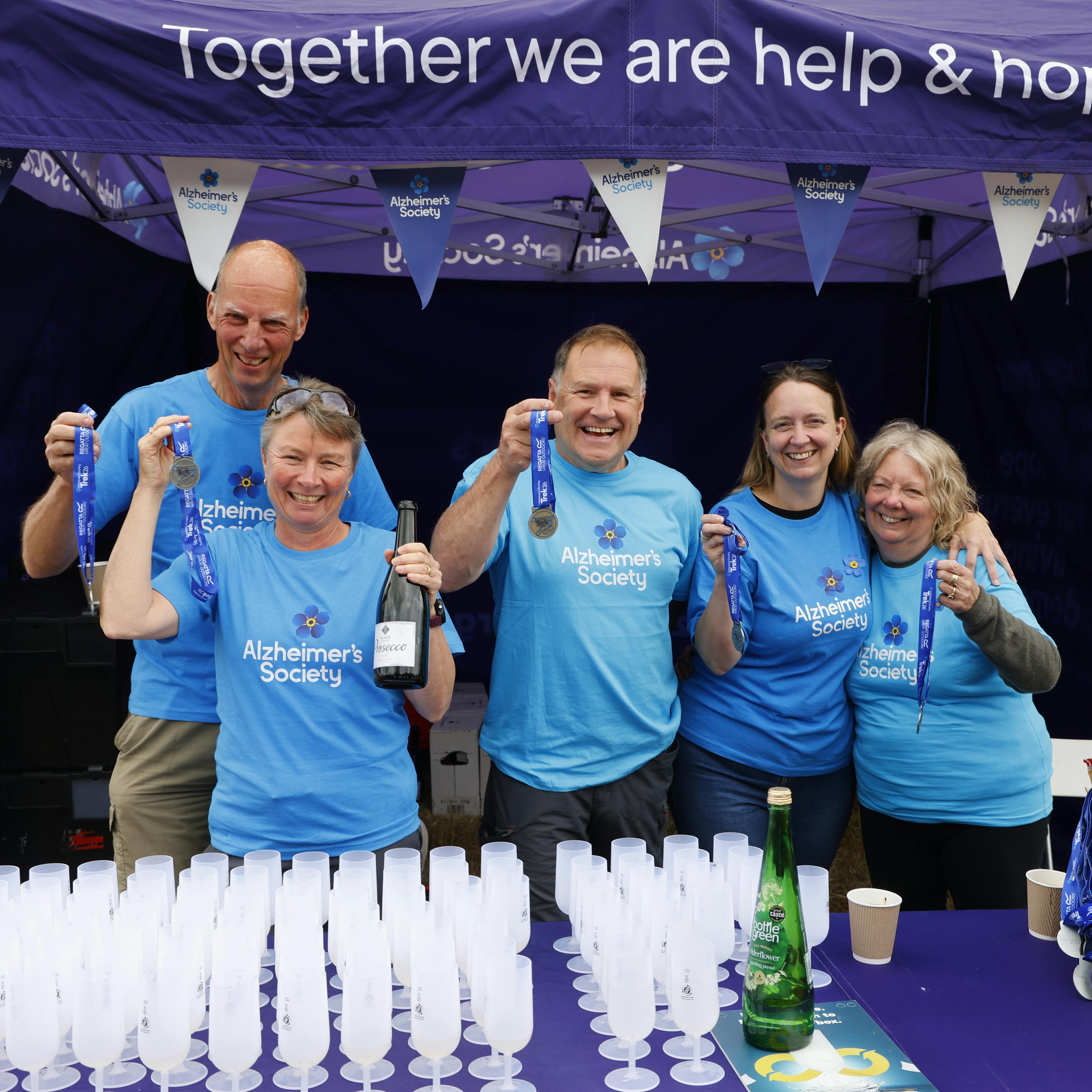 Five Trek26 volunteers standing behind a table filled with prosecco flutes and holding up medals.