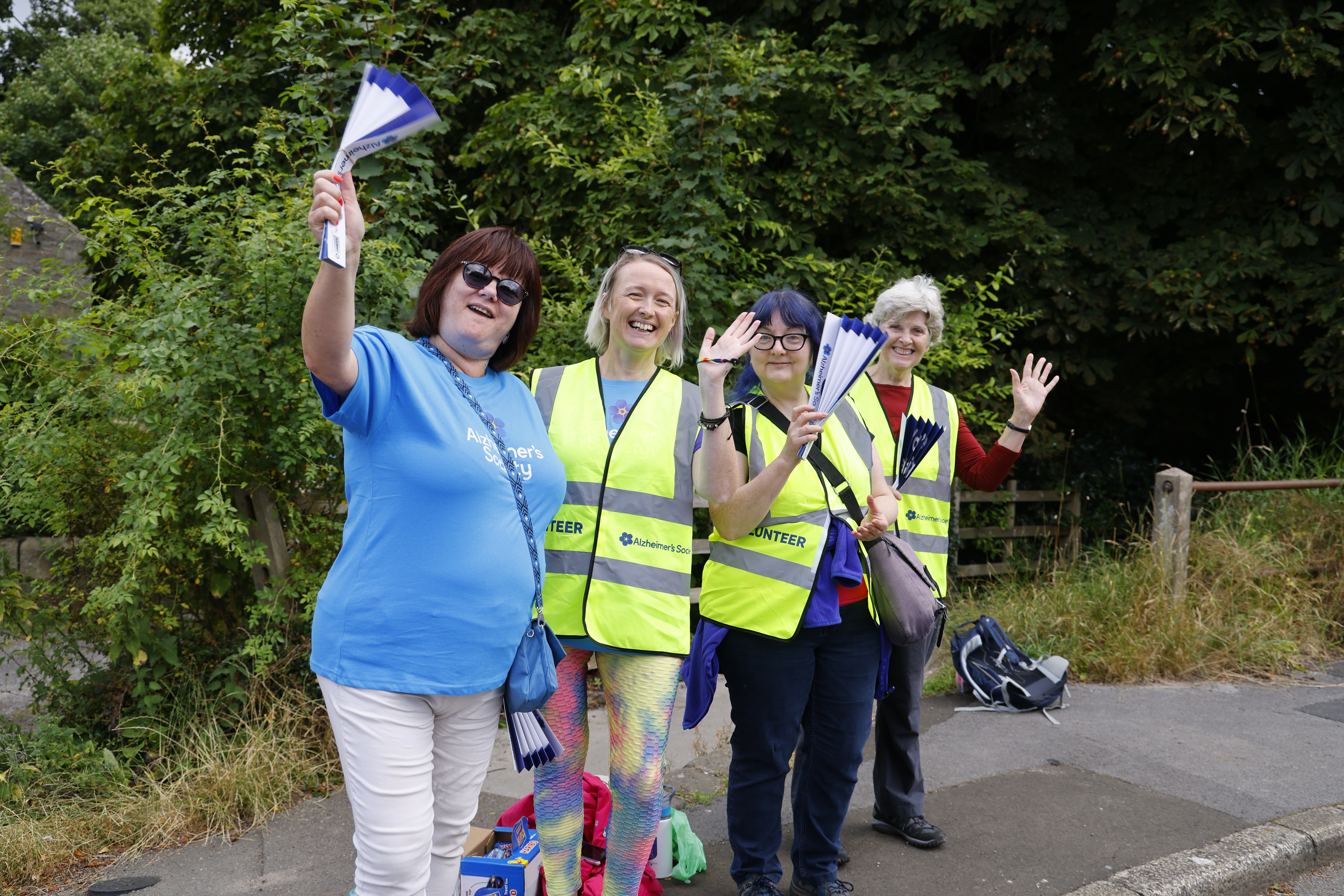Four Trek26 volunteers standing on the roadside, wearing high vis vests and waving.
