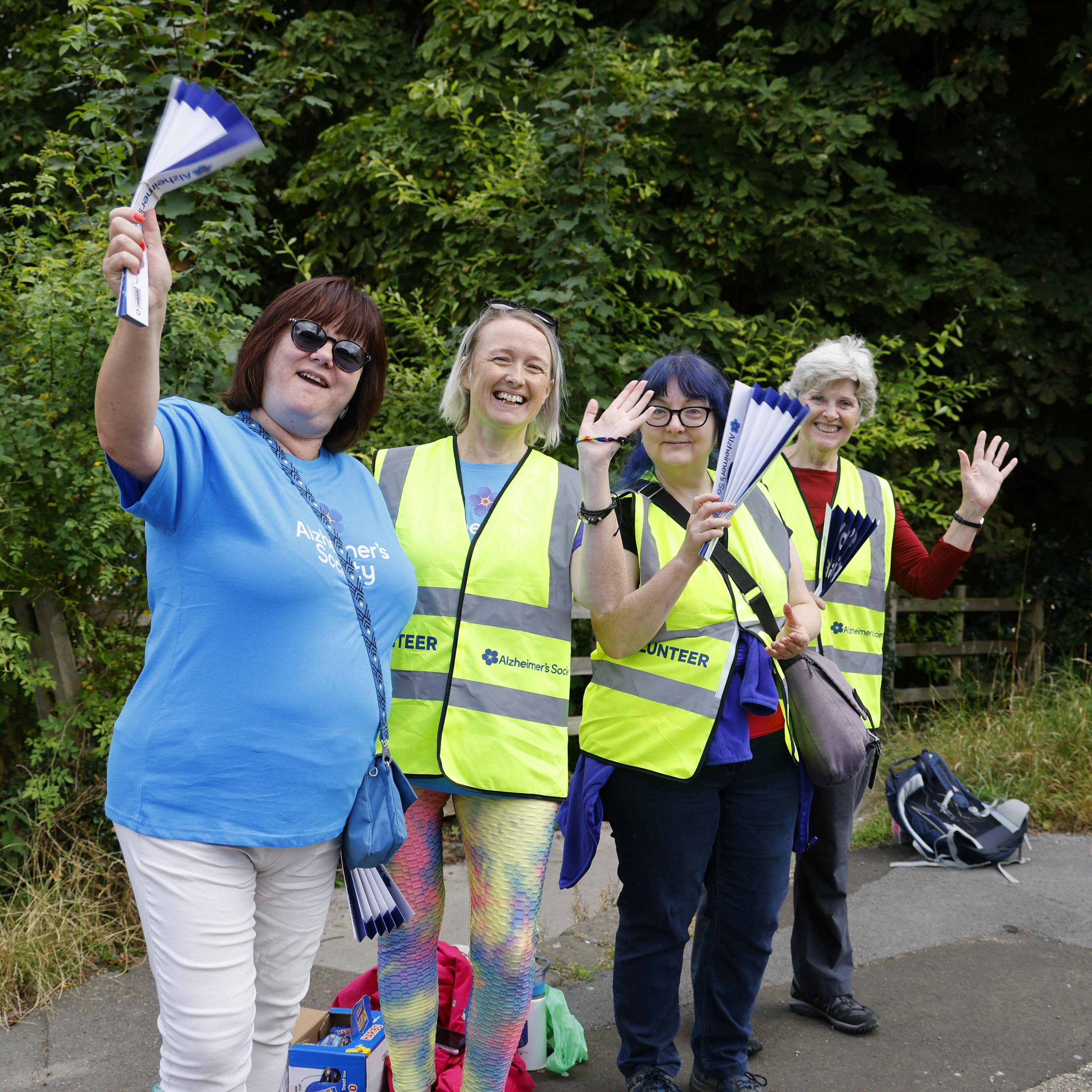 Four Trek26 volunteers standing on the roadside, wearing high vis vests and waving.