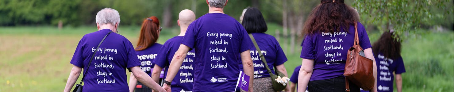 A group of people walking away from the camera, all wearing Alzheimer Scotland t-shirts. The message on the back of each shirt reads: 'Every penny raised in Scotland stays in Scotland.'