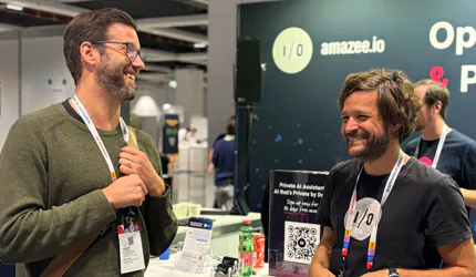 Two smiling tech professionals wearing event lanyards are conversing at an amazee.io exhibition booth showcasing open source cloud hosting and private AI solutions.