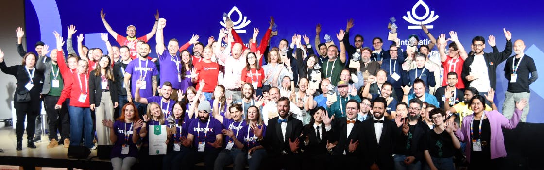 A large group of people celebrating on a stage with their hands raised, at the Splash Awards International ceremony. The background is a bright blue screen featuring a white, stylized flame-drop logo (similar to the Drupal logo). Many people are wearing t-shirts and holding awards. The photo captures a moment of collective excitement and celebration.
