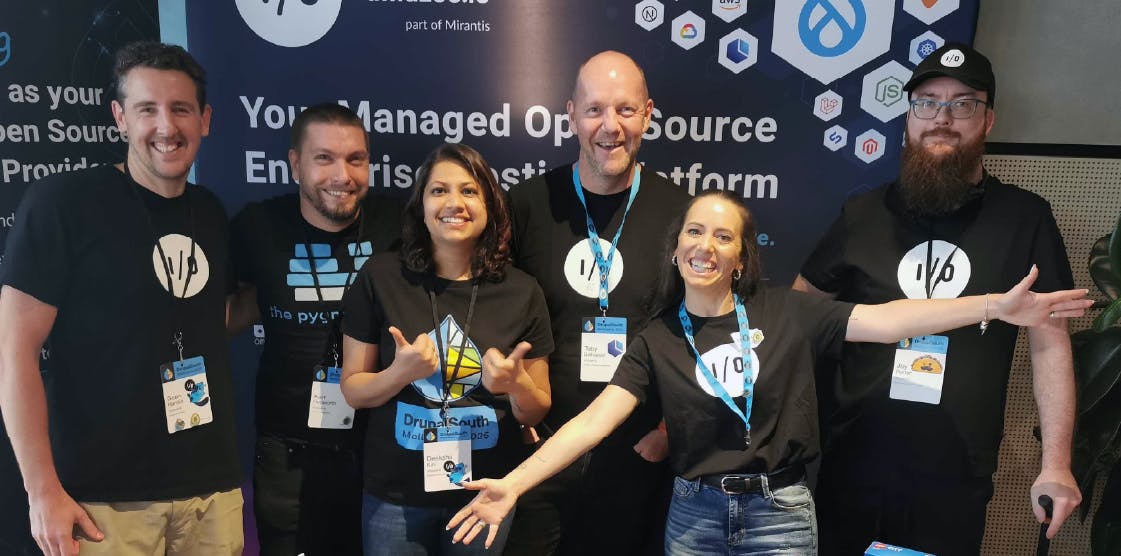 Six members of the amazee.io team smiling together in front of a company banner at the DrupalSouth conference. They are wearing black branded t-shirts with the I/O logo.