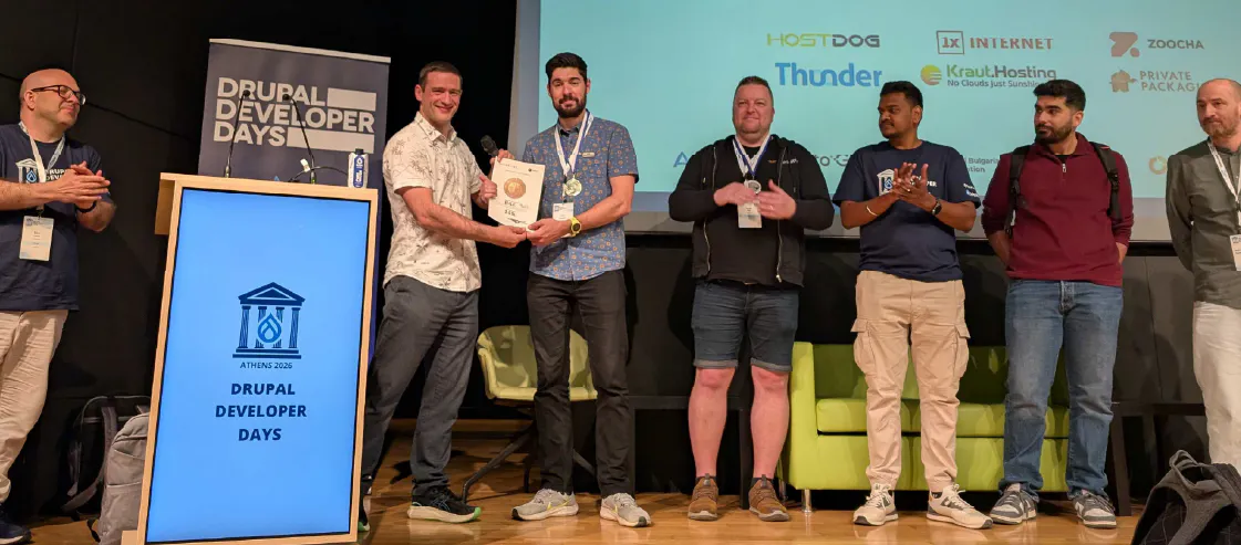 An awards ceremony on stage at Drupal Developer Days Athens 2026. Several participants are standing together, with one individual receiving a certificate while others applaud. A digital podium in the foreground displays the event logo and name, while the background screen shows various sponsor logos, including amazee.io.