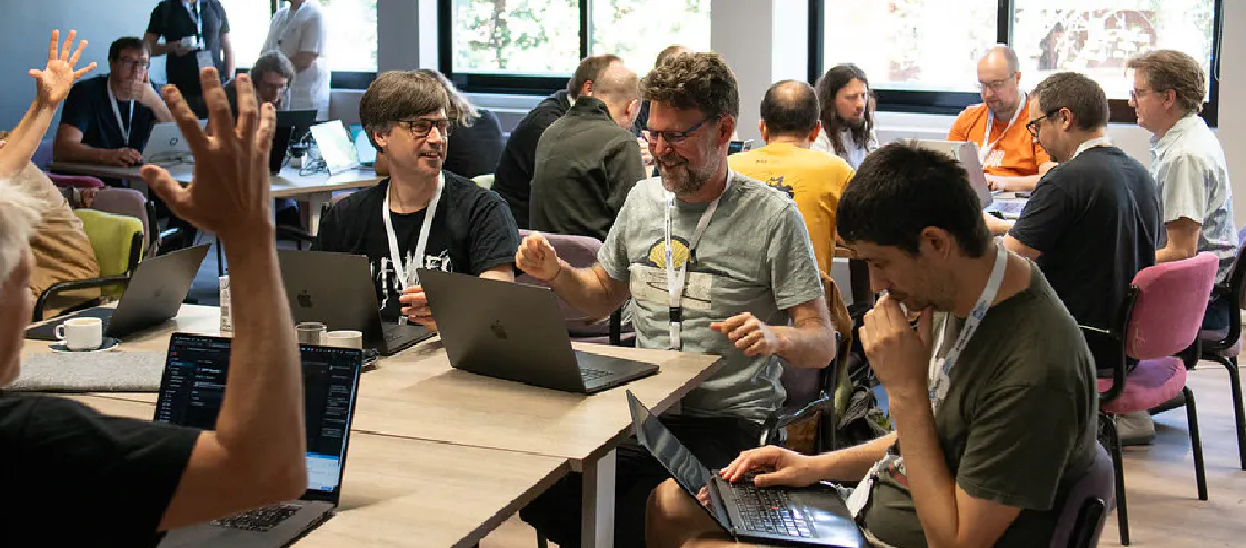 A group of developers working in collaborative groups at Drupal Developer Days Athens 2026. People are seated at long tables with laptops, engaged in discussion and coding, representing a community contribution sprint or workshop in a bright, modern event space.
