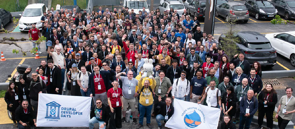A large group of attendees posing for a photo outdoors at Drupal Developer Days Athens 2026. Two people in the foreground are holding large banners: one for the event itself and another for the Greek Drupal Community. Many participants are waving, and a plush mascot is held up in the center of the crowd.