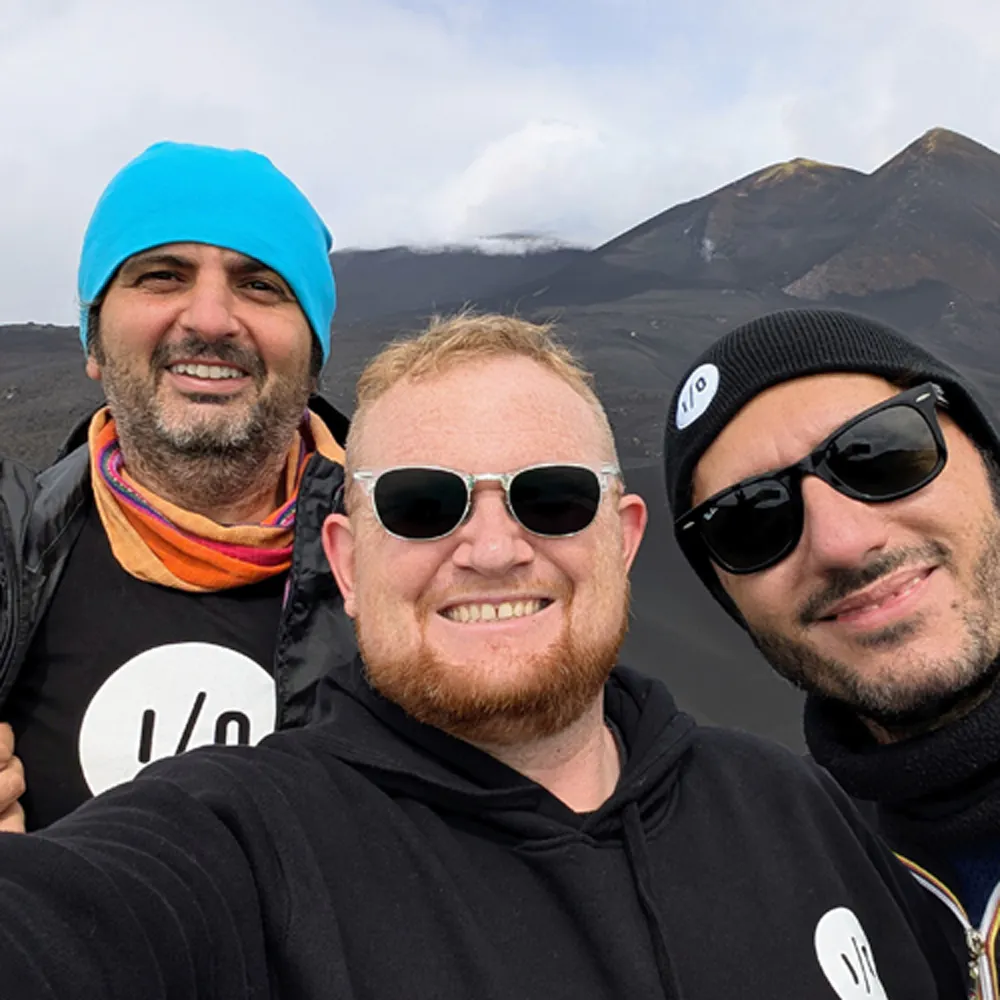amazee.io engineering team members take an outdoor selfie on Mount Etna, wearing branded black hoodies, beanies, and i/o t-shirts during a get-together in Sicily.