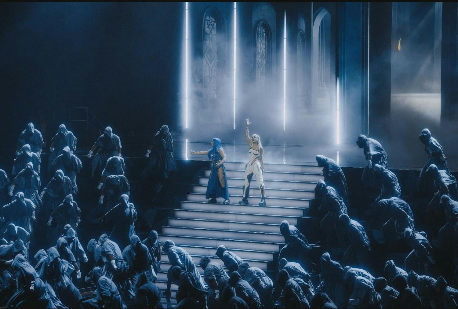 Two people performing on a stage surrounded by hooded figures