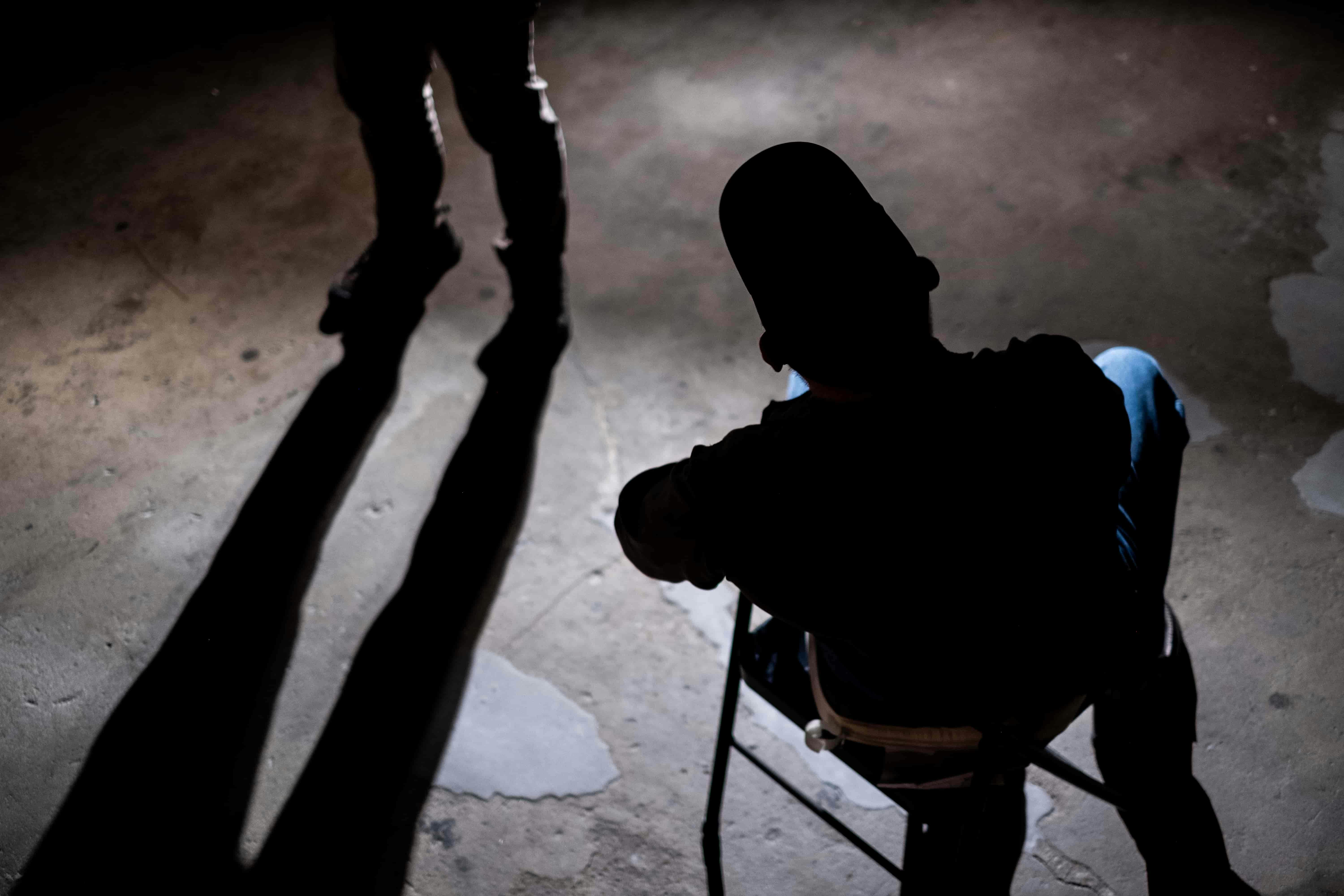 Dark figure of man sat on chair with legs of someone stood next to him