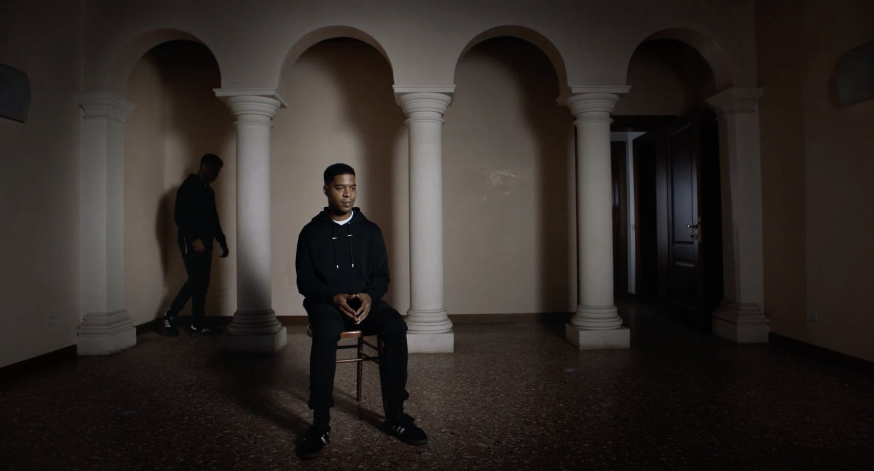 Man sat on chair in room with figure walking behind him