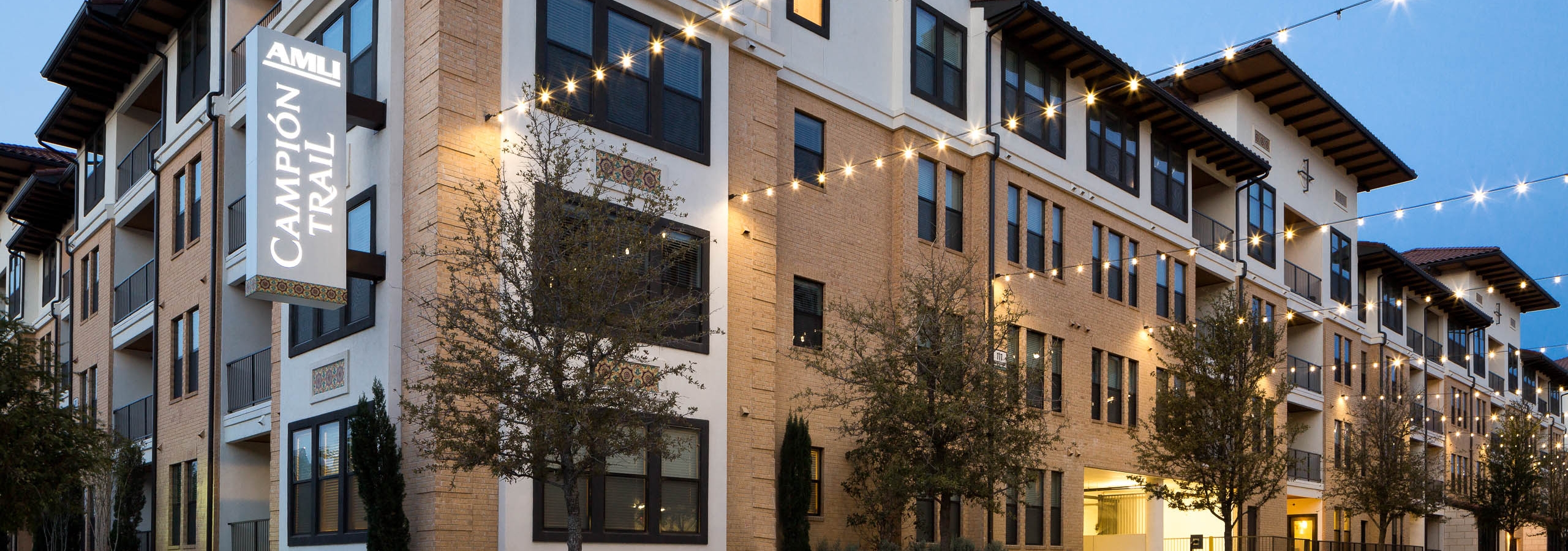 Nighttime exterior of AMLI Campion Trail apartment building brick facade with lit building sign and trees and string lights