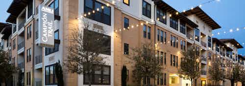 Nighttime exterior of AMLI Campion Trail apartment building brick facade with lit building sign and trees and string lights