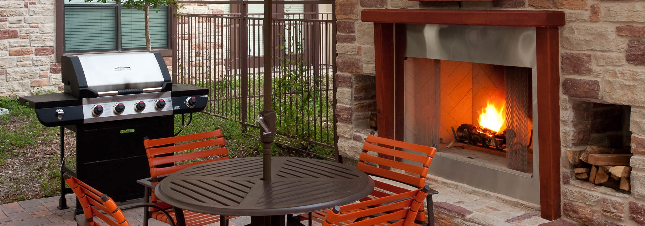Daytime view of AMLI at Escena brick outdoor fireplace with brown table and four orange chairs and grill