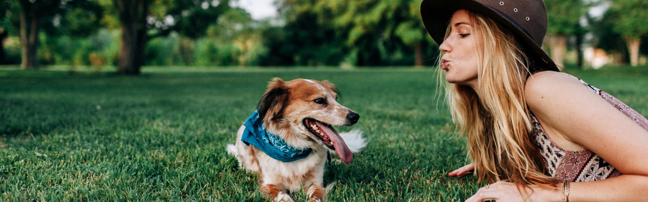 Blonde girl with brown hat with dog laying in green grass at park with larger trees blurred in the background