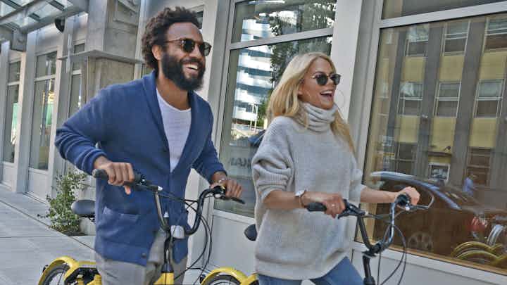 Man and woman wearing sunglasses and smiling while riding their bikes through town and stopping in front of a storefront
