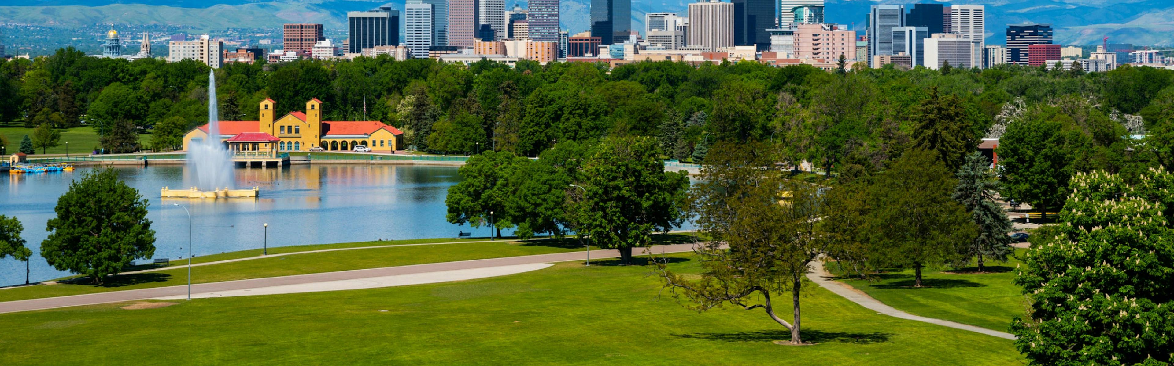 A view of City Park with a lake and surrounding trees and a view of the downtown Denver skyline and a mountain background