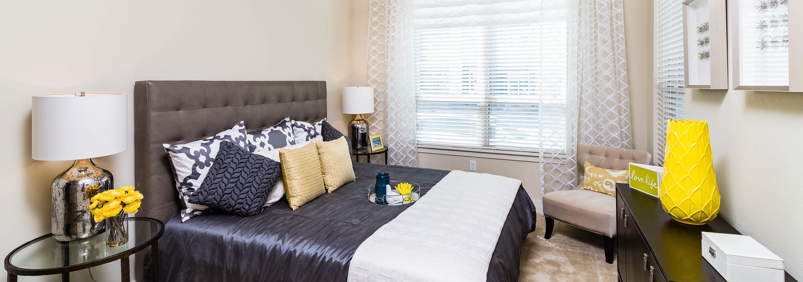 Interior view of AMLI RidgeGate bedroom with queen bed and gray upholstered headboard facing large window with sheer curtains