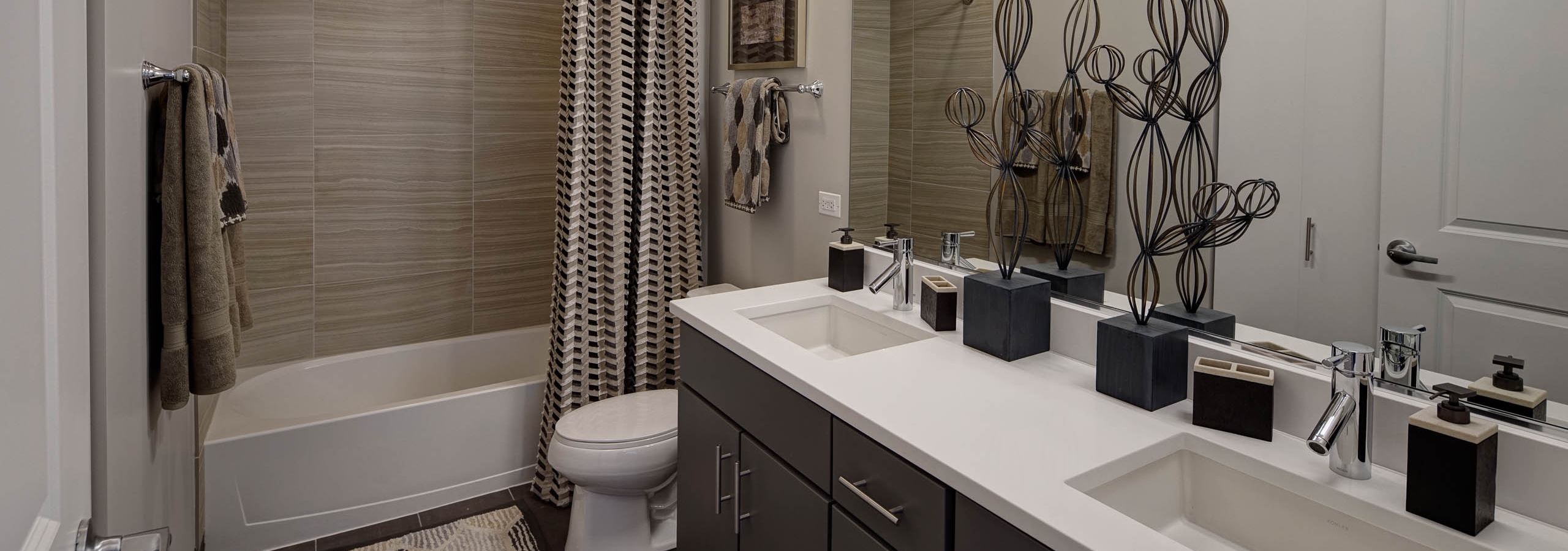 Interior view of bathroom at AMLI Lofts containing a double vanity, toilet and bathtub with a striped curtain and beige tiles