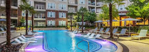 Nighttime view of AMLI Frisco Crossing apartments swimming pool area with lounge seating, umbrellas and palm trees