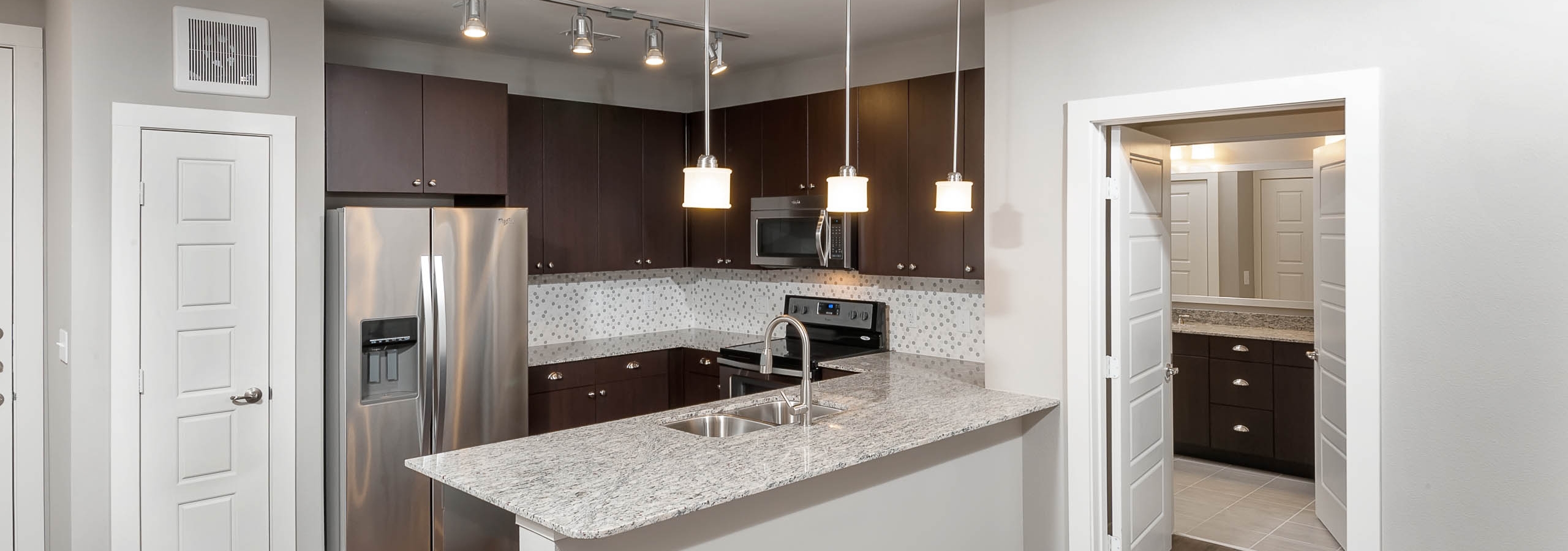 Interior of AMLI Frisco Crossing apartment kitchen with espresso cabinetry and tile backsplash and stainless steel appliances