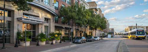 Exterior view of AMLI Las Colinas apartment building with cars parked and planters in front and DART train passing at right